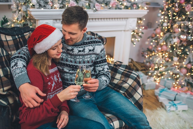 Hermosa pareja se sienta junto y sostiene copas de champaña. Joven abrazo mujer. Ella mira las gafas. Se sientan en una habitación decorada.