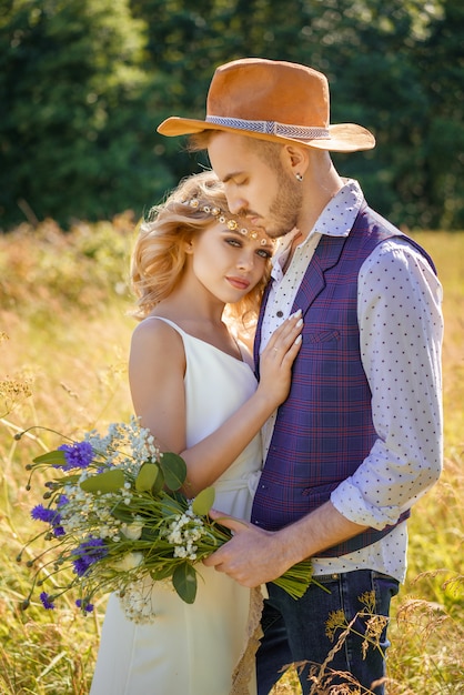 Hermosa pareja con un sombrero en el campo abrazando en el verano en el campo. El chico tiene un ramo de flores. Relacion romantica