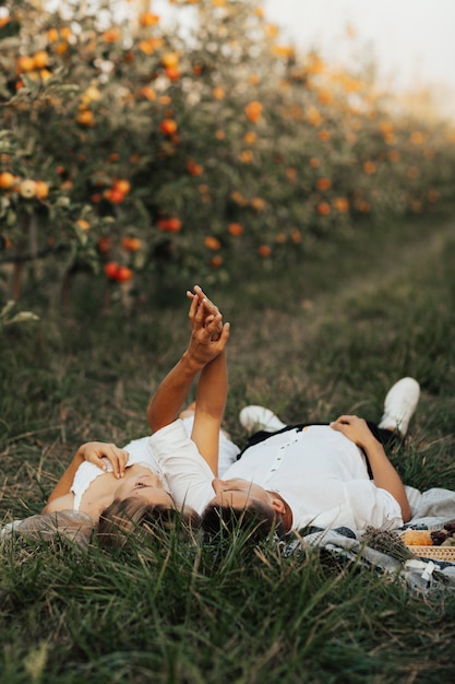 Hermosa pareja sonriente disfrutando de un día de picnic en el huerto de manzanas. Ellos acostados y tomados de la mano.