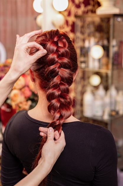 Hermosa pelirroja con cabello largo, peluquera teje una trenza francesa, en un salón de belleza.