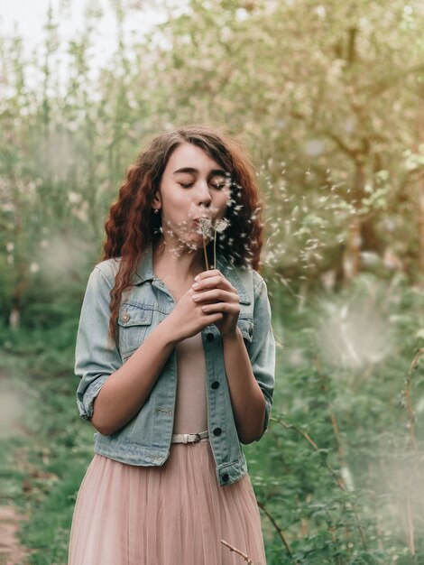 Foto hermosa pelirroja rizada niña soplando diente de león en un jardín de primavera