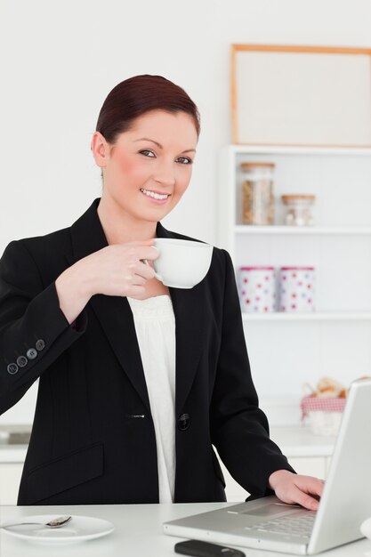 Hermosa pelirroja en traje relajante con su computadora portátil mientras desayunando en la cocina