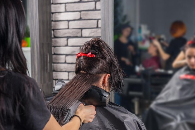 Hermosa peluquería peinando y usando secador de pelo en el cabello de la joven morena mientras ella está sentada en un sillón en el salón de belleza