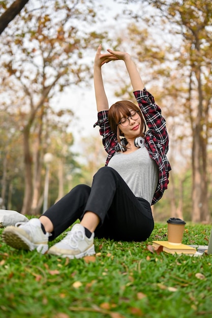 Foto hermosa y relajada joven asiática estirando las manos sentada en la hierba en el parque