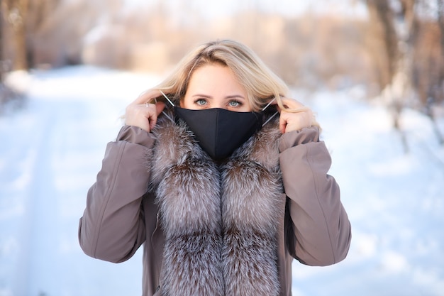 Una hermosa rubia con un abrigo de piel con cuello de piel se pone una máscara protectora negra