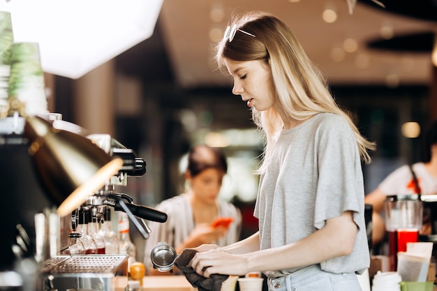 Una hermosa rubia delgada con cabello largo, vestida con un atuendo informal, está cocinando café en una cafetería moderna. Se muestra el proceso de elaboración del café. .