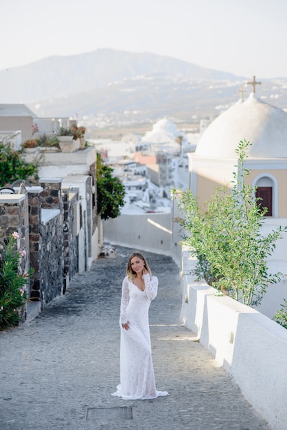 Hermosa rubia delgada mujer top model novia con cabello largo y piernas en el vestido de novia blanco con un largo tren y volantes se encuentra en una pintoresca isla de Santorini en Grecia