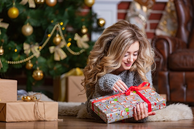 Hermosa rubia se recuesta sobre una alfombra cálida y mira un brillante regalo de Navidad con una cinta roja cerca del árbol de Navidad