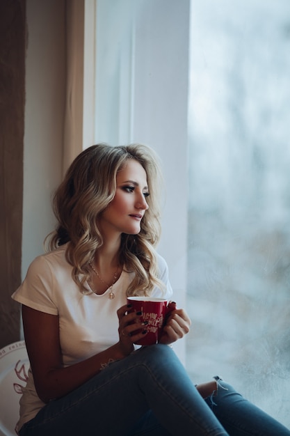 Hermosa rubia sentada en el alféizar de la ventana con taza