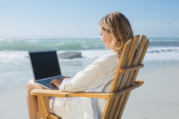 Hermosa rubia sentada en la silla de cubierta usando la computadora portátil en la playa