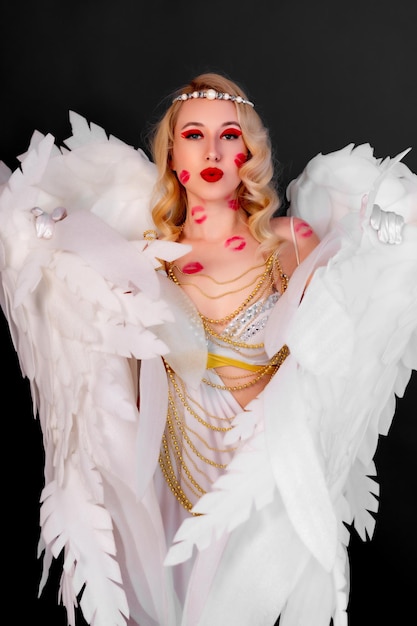 Hermosa rubia con un sexy vestido blanco decorado con cuentas doradas y grandes alas blancas en la espalda Rastros de besos en el cuerpo y la cara Cupido sexy Fondo negro