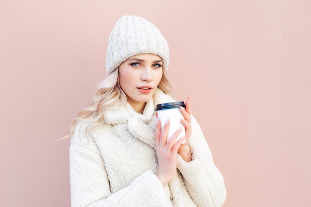 Hermosa rubia con un sombrero blanco y abrigo sostiene una taza de café