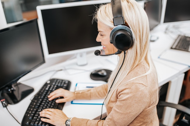 Hermosa rubia sonriente agente de centro de llamadas con auriculares trabajando en soporte en la oficina.