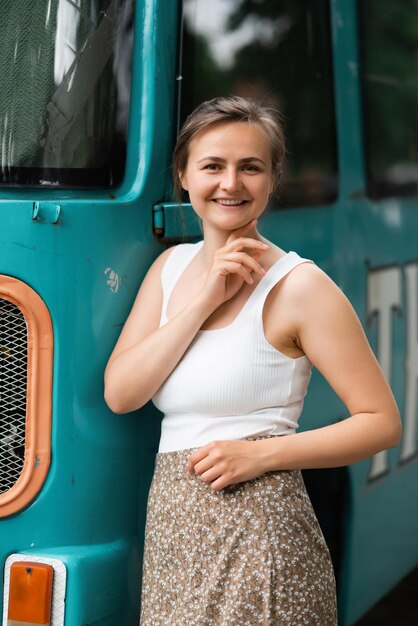 Una hermosa rubia sonriente está parada al lado de un viejo autobús escolar y mirando a la cámara