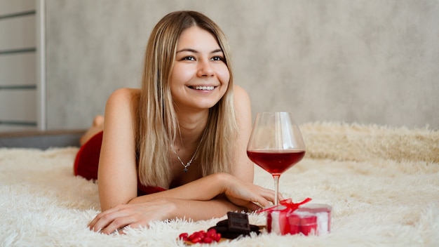 Una hermosa rubia sonriente yace en la cama. Mañana del día de San Valentín. Una copa de vino, chocolate, dulces y un obsequio junto a la niña. Feliz mañana en el amor