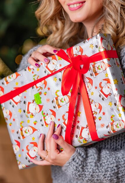 Hermosa rubia sosteniendo un brillante regalo de Navidad con cinta roja cerca del árbol de Navidad Vista recortada