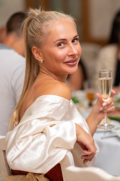 Hermosa rubia con un traje blanco se sienta en un restaurante con una copa de champán.