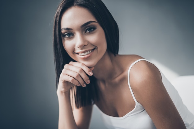 Hermosa seductora. Atractiva mujer joven sosteniendo la mano en la barbilla y mirando a la cámara con una sonrisa mientras está sentado contra la pared gris