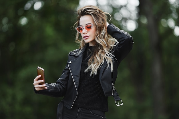 Hermosa y sensual chica rubia modelo con chaqueta de cuero negro, jeans y elegantes gafas de sol toma una selfie en su teléfono inteligente y posa al aire libre en la calle de la ciudad