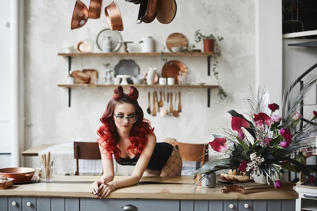 Hermosa y sexy chica modelo pin-up con cabello rojo y maquillaje profesional, en monos negros y gafas posando en una cocina.