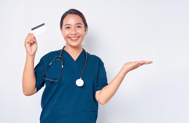 Hermosa y sonriente enfermera profesional asiática joven que trabaja con uniforme azul sosteniendo una tarjeta de crédito y mostrando un espacio vacío con la palma aislada en el fondo blanco Concepto de medicina sanitaria