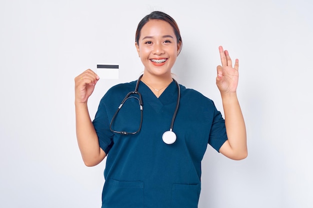 Hermosa y sonriente enfermera profesional asiática joven que trabaja con uniforme azul sosteniendo una tarjeta de crédito y mostrando un gesto correcto aislado en el fondo blanco Concepto de medicina de la salud