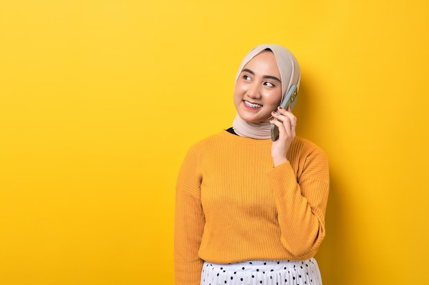 Hermosa sonriente feliz chica asiática usando hiyab hablando por teléfono móvil con un amigo mirando el espacio vacío aislado en el fondo amarillo