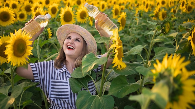 Hermosa y sonriente granjera de mediana edad sosteniendo botellas de aceite de girasol dorado en sus manos en un campo de cosecha de girasoles en un día soleado