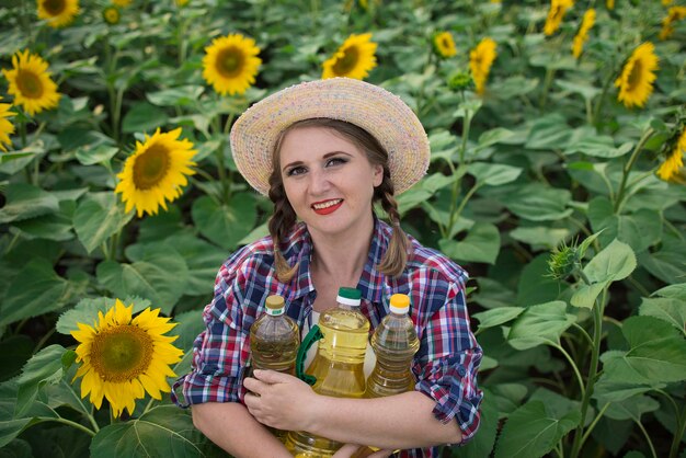 Hermosa y sonriente granjera de mediana edad sosteniendo botellas de aceite de girasol dorado en sus manos en un campo de cosecha de girasoles en un día soleado