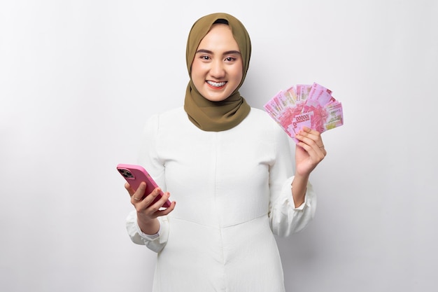 Hermosa sonriente joven árabe asiática musulmana con hiyab sosteniendo dinero en efectivo en billetes de rupias y teléfono móvil aislado en fondo blanco Concepto de estilo de vida religioso de la gente