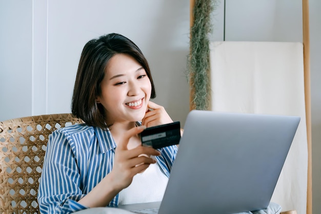 Una hermosa y sonriente joven asiática sentada en un sillón de ratán en la sala de estar de su casa administrando la banca en línea con su portátil y tarjeta de crédito La tecnología hace la vida mucho más fácil
