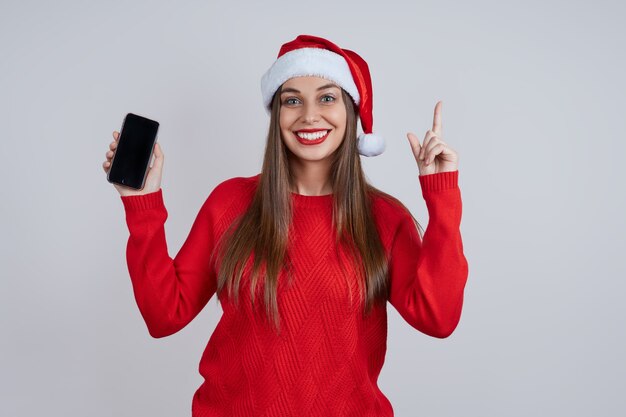 Una hermosa y sonriente joven con un gorro de Papá Noel, con un teléfono en la mano, señala con el dedo la pantalla. Concepto de compras online, saludos online, descuentos navideños.