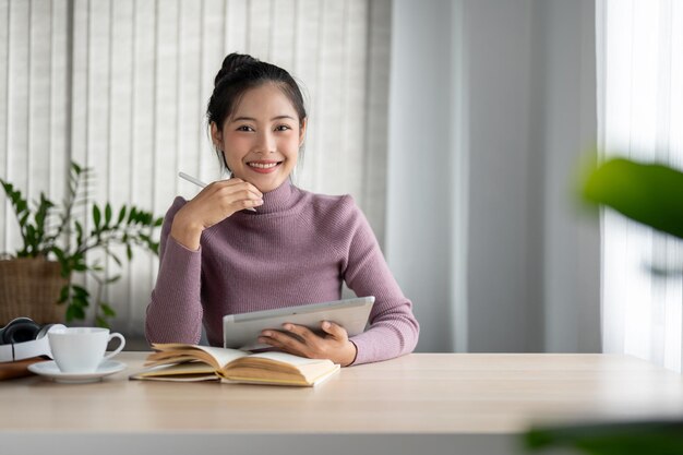 Una hermosa y sonriente mujer asiática sentada en una mesa en su sala de estar con su tableta digital