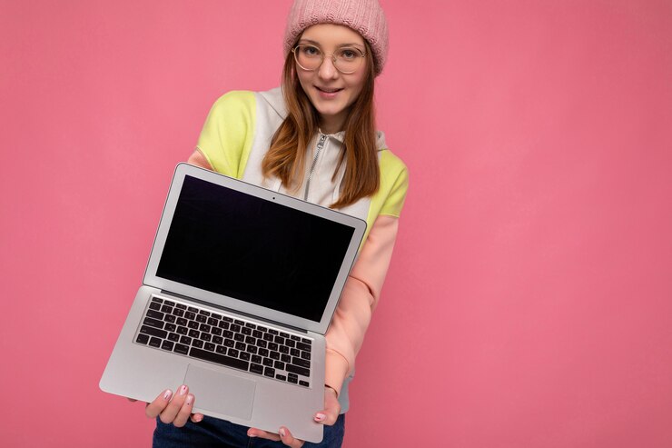 Foto hermosa sonriente mujer joven y bonita con sombrero colorido suéter y gafas sosteniendo computadora portátil con monitor de pantalla de espacio de copia vacía con auriculares blancos mirando a la cámara de pie aislado