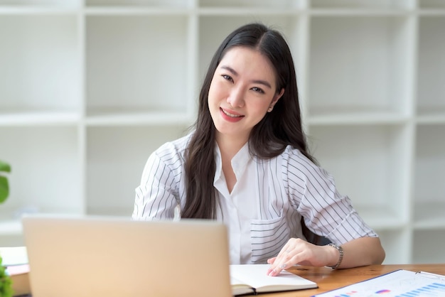 Una hermosa y sonriente mujer de negocios asiática sentada con una laptop y una computadora mientras hacía papeleo en la oficina. Mirando a la cámara.