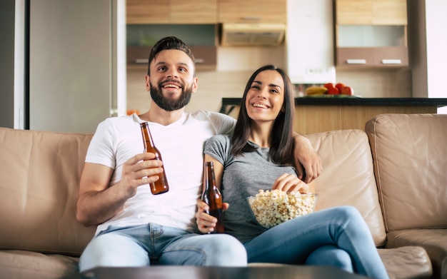 Hermosa y sonriente pareja moderna enamorada está viendo una película o una película en la televisión mientras se relajan en el sofá de su propio apartamento