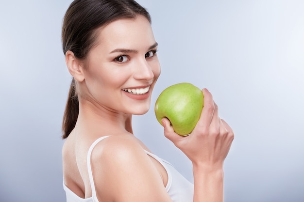 Hermosa sonrisa, dientes blancos y fuertes. Cabeza y hombros de mujer joven con sonrisa blanca como la nieve con manzana verde, cuidado de los dientes. Se apartó un poco