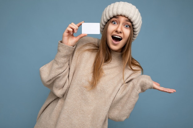 Hermosa sorprendida positiva sonriente joven rubia oscura vestida con suéter beige y sombrero beige tejido aislado sobre fondo azul sosteniendo y mostrando la tarjeta de crédito mirando a la cámara.