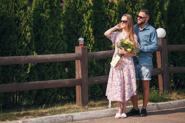 Una hermosa y tierna mujer con cabello rubio, un vestido ligero y un ramo de flores camina en un parque soleado con su guapo novio con una camisa azul y pantalones cortos.