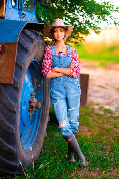 Hermosa vaquera rubia con sombrero de paja y mezclilla de pie en general cerca del tractor azul.