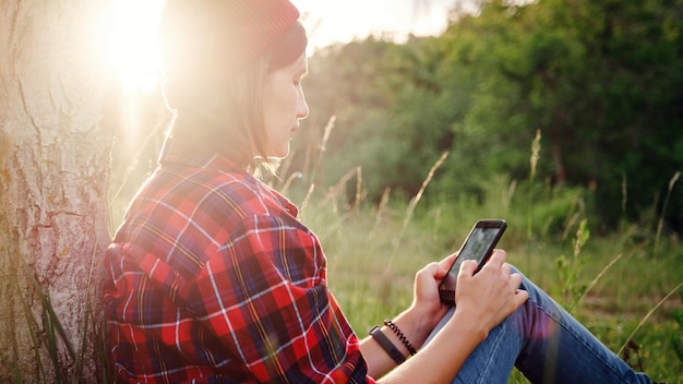 Hermosa viajera sentada cerca del árbol y escribiendo en el teléfono móvil