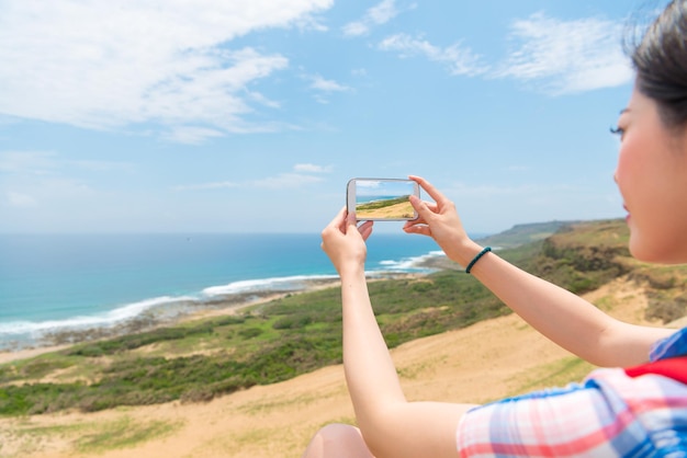 hermosa viajera sonriente en la cima de la montaña usando un teléfono móvil tomando fotos espectaculares del paisaje marino a través de la aplicación de tecnología en vacaciones.