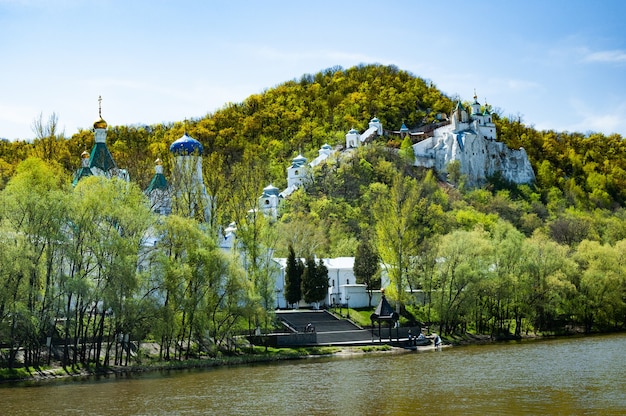 Hermosa vista hechizante de la Iglesia Ortodoxa y el monasterio ubicado a orillas del río en las colinas cubiertas de matorrales verdes en un soleado día de otoño. Concepto de vida espiritual