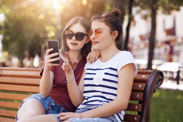 Hermosas adolescentes en tonos de moda, miran atentamente el teléfono inteligente, hacen compras en línea, eligen un nuevo atuendo de verano, posan en el banco en el parque, disfrutan del clima cálido y soleado. Concepto de recreación