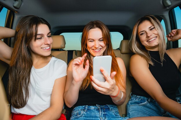 Hermosas amigas se divierten, sonriendo con el teléfono Mobiel en el respaldo del asiento del vehículo, dentro del salón del automóvil