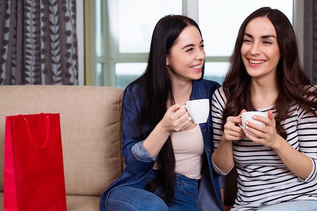 Hermosas amigas sonrientes con ropa informal sentadas en el sofá de casa hablando y bebiendo café o té