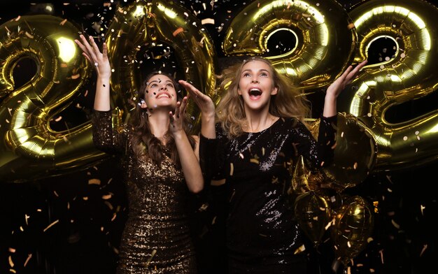 Hermosas chicas felices con elegantes vestidos de fiesta sexy con globos de oro 2022, divirtiéndose en la fiesta de Navidad o Año Nuevo.