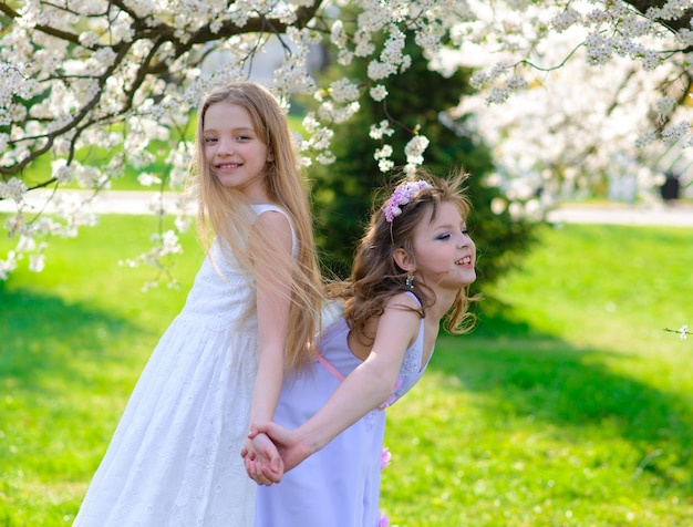 Hermosas chicas jóvenes con ojos azules en vestidos blancos en el jardín con manzanos floreciendo divirtiéndose