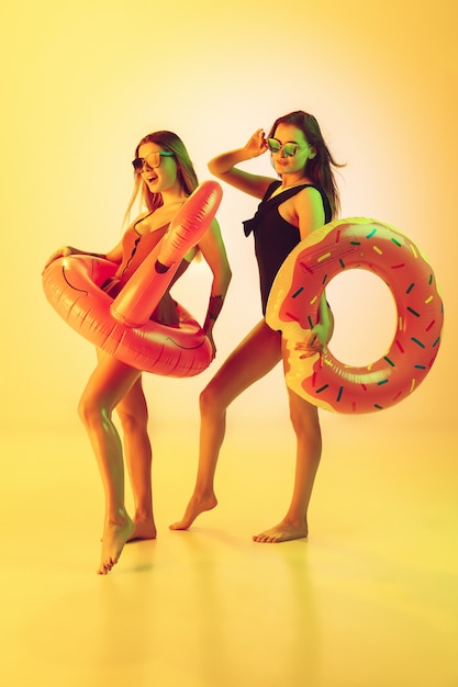 Hermosas chicas en trajes de baño de moda aislados sobre fondo amarillo de estudio en luz de neón. Concepto de verano, resort, moda y fin de semana. Posando en rosquilla de goma y flamenco con emociones brillantes.