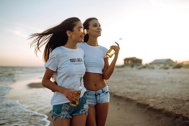 Hermosas dos chicas con gafas sostienen una botella con cerveza en sus manos en la playa Estilo de vida de verano